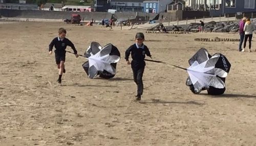 Dau blentyn yn rhedeg ar hyd traeth Pentywyn gyda pharasiwtiau bach yn llusgo ar eu hôl