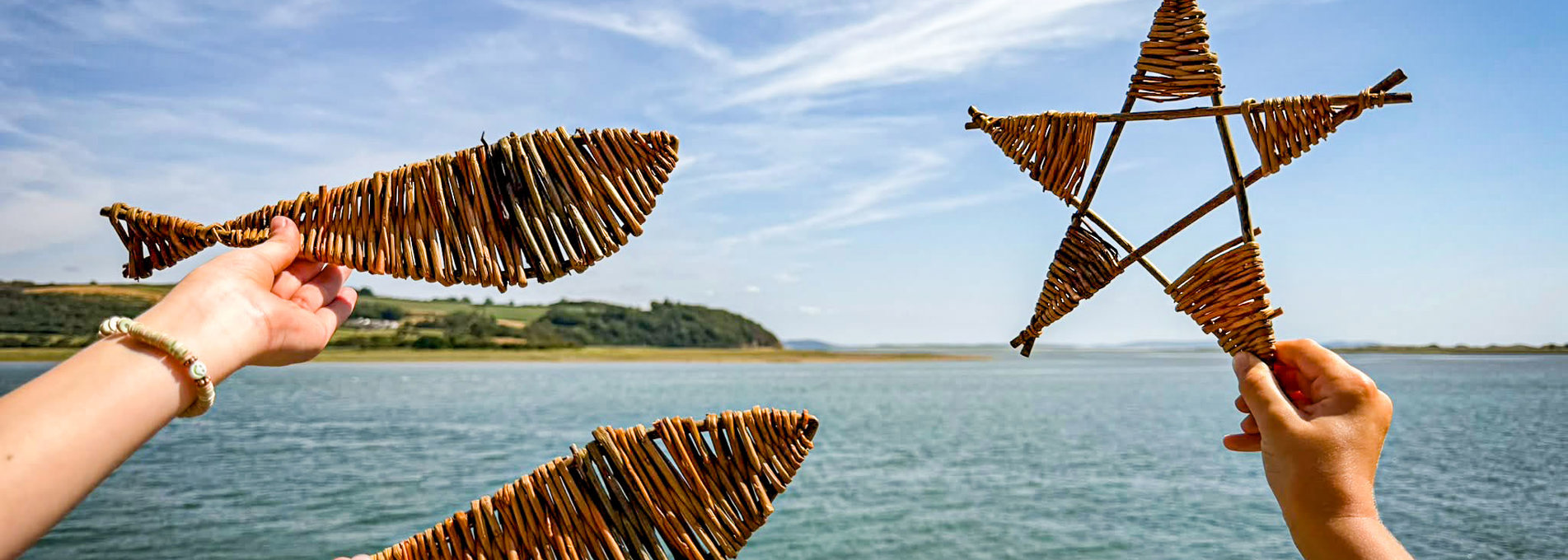 Dwylo yn dal pysgod helyg wedi'u gwneud â llaw ac addurniadau gwehyddu siâp seren yn erbyn cefndir arfordirol, gyda dyfroedd aberoedd a glannau i'w gweld yn y pellter
