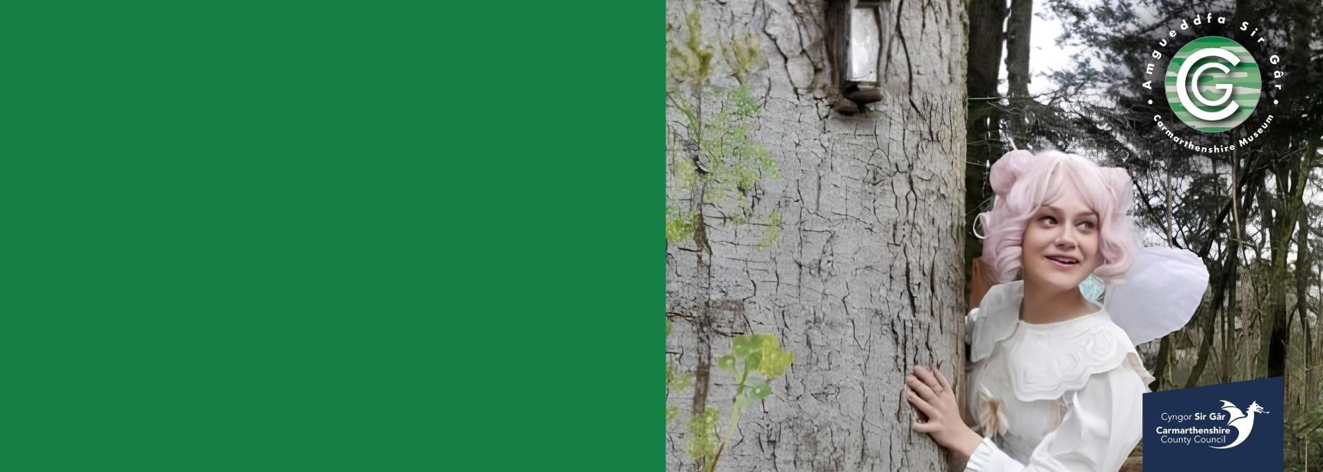 Menyw wedi'i gwisgo fel tylwyth teg yn edrych allan o'r tu ôl i goeden