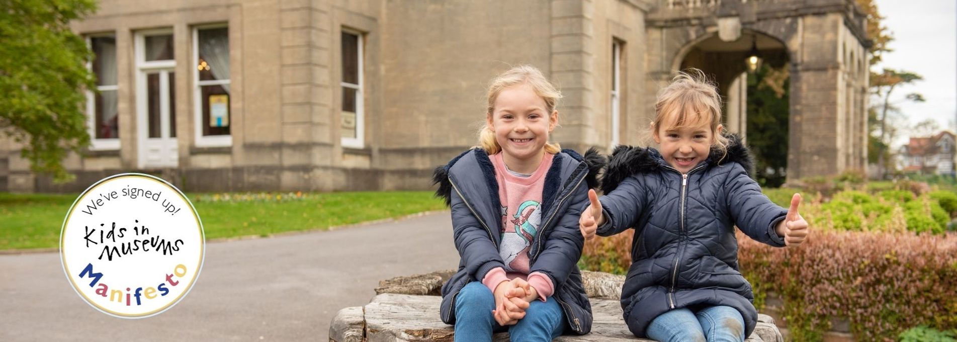 Dau blentyn yn gwenu yn eistedd ar fonyn coeden o flaen Amgueddfa Parc Howard